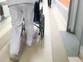 Osoba w białym ubraniu prowadzi wózek inwalidzki korytarzem szpitalnym.