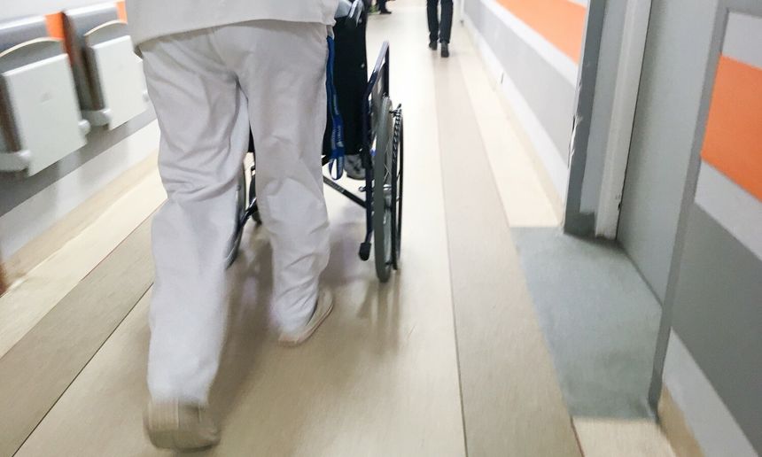 Osoba w białym ubraniu prowadzi wózek inwalidzki korytarzem szpitalnym.