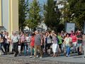 Grupa osób spacerujących po ulicy, w tle drzewa i budynki. Słoneczna pogoda.