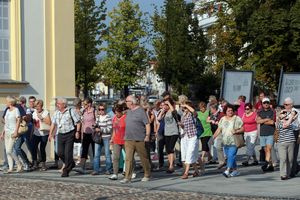 Grupa osób spacerujących po ulicy, w tle drzewa i budynki. Słoneczna pogoda.