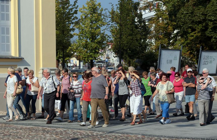 Grupa osób spacerujących po ulicy, w tle drzewa i budynki. Słoneczna pogoda.