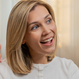 Kobieta z blond włosami uśmiecha się i gestykuluje, siedząc w jasnym wnętrzu.