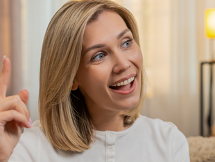 Kobieta z blond włosami uśmiecha się i gestykuluje, siedząc w jasnym wnętrzu.