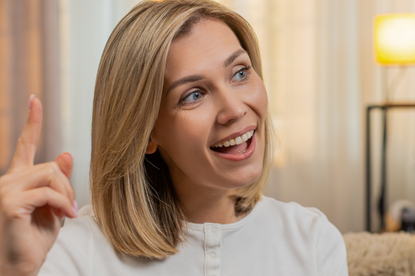 Kobieta z blond włosami uśmiecha się i gestykuluje, siedząc w jasnym wnętrzu.