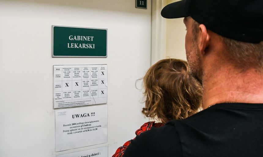 Rodzina stoi przed drzwiami gabinetu lekarskiego, widoczna informacja o terminach wizyt.