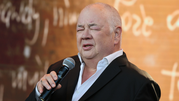 Mężczyzna w garniturze trzyma mikrofon, wyraża emocje podczas wystąpienia publicznego.
