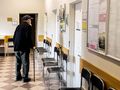 Mężczyzna w kapeluszu z laską stoi w korytarzu z krzesłami i zamkniętymi drzwiami.