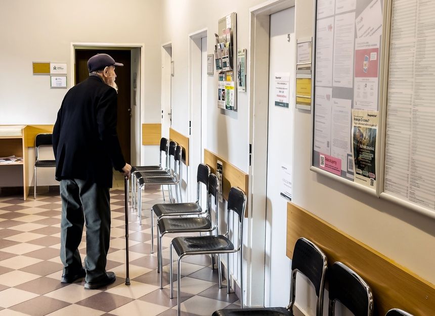 Mężczyzna w kapeluszu z laską stoi w korytarzu z krzesłami i zamkniętymi drzwiami.