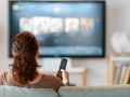 Kobieta siedzi na kanapie z pilotem w ręku, oglądając telewizję w jasnym salonie.