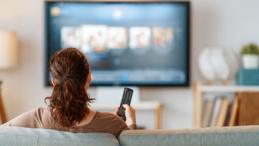 Kobieta siedzi na kanapie z pilotem w ręku, oglądając telewizję w jasnym salonie.