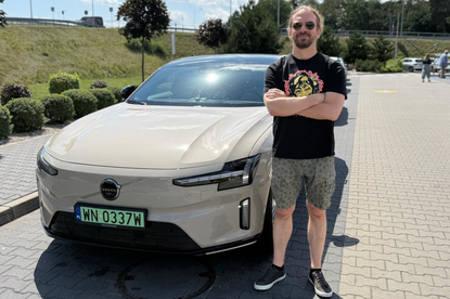 Mężczyzna stoi obok kremowego samochodu Volvo na parkingu, ma na sobie czarną koszulkę i okulary.