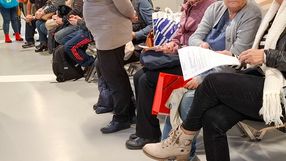 Grupa osób siedzących na ławkach, czekających w kolejce. Widać torby i dokumenty.