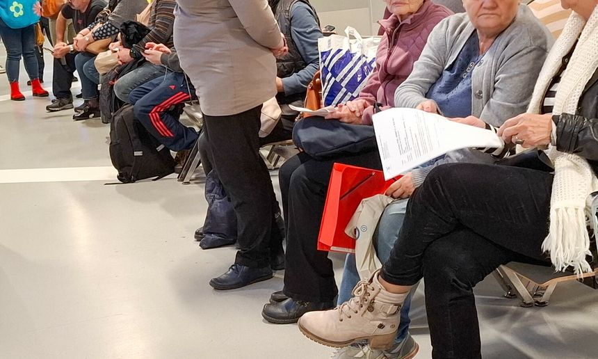 Grupa osób siedzących na ławkach, czekających w kolejce. Widać torby i dokumenty.