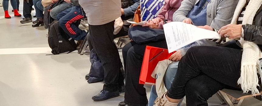 Grupa osób siedzących na ławkach, czekających w kolejce. Widać torby i dokumenty.