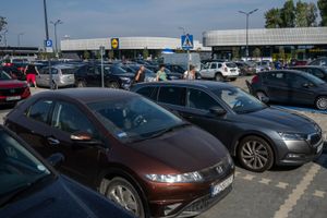 Zdjęcie przedstawia zatłoczony parking przy sklepie Lidl, z wieloma samochodami i przechodzącymi ludźmi.