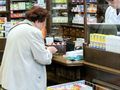 W aptece, starsza kobieta przegląda leki, za ladą stoi farmaceuta w białym fartuchu.