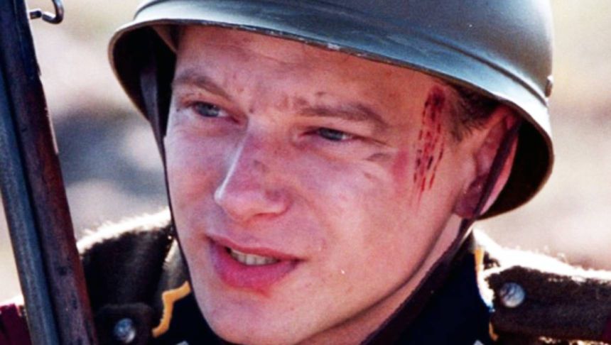 Mężczyzna w hełmie wojskowym z zadrapaniem na twarzy, trzymający karabin.