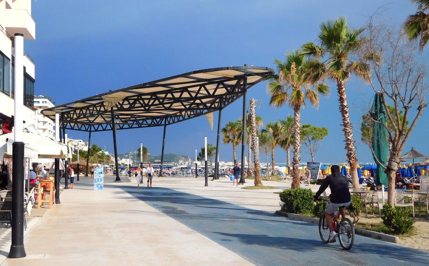 Promenada nadmorska z palmami, rowerzystą i turystami. W tle plaża i chmury na niebie.