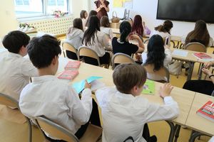 Uczniowie w białych koszulach siedzą w klasie, słuchając nauczyciela przy tablicy.
