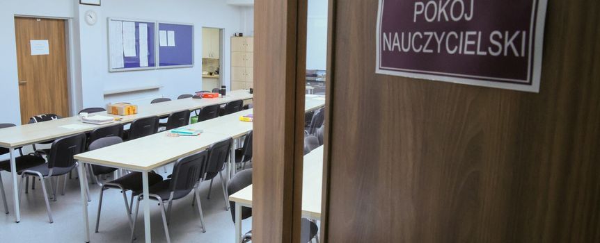 Wnętrze pokoju nauczycielskiego z wieloma stołami i krzesłami, widok przez otwarte drzwi.