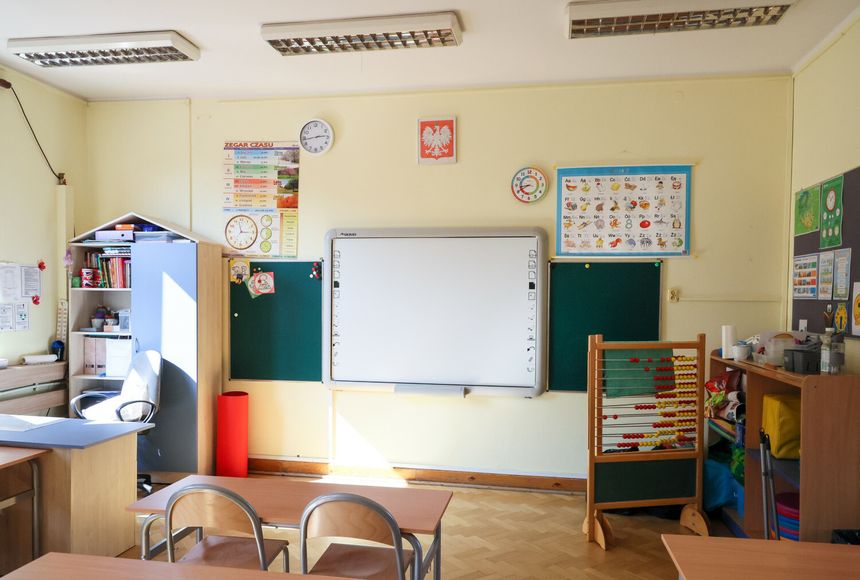 Klasa szkolna z tablicą, biurkiem nauczyciela i materiałami edukacyjnymi.