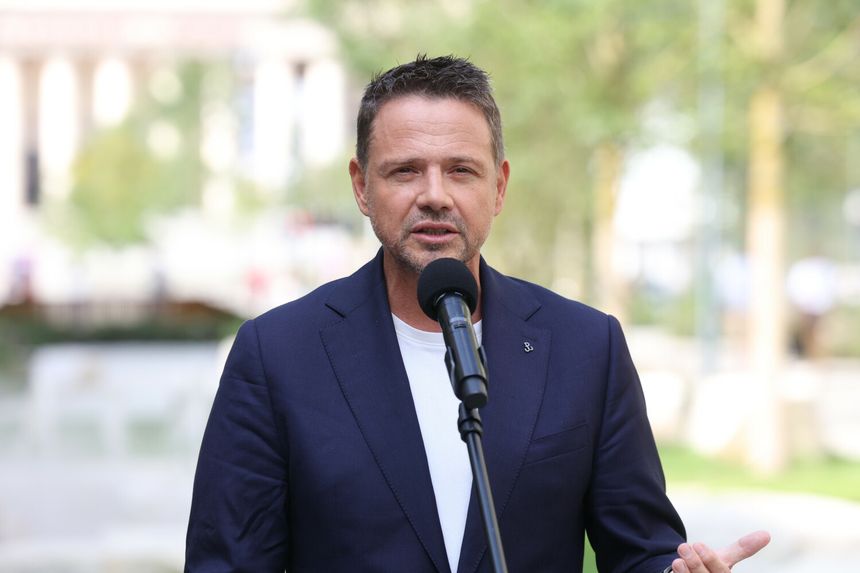 Mężczyzna w garniturze przemawia do mikrofonu w parku. Zielone tło, słoneczny dzień.