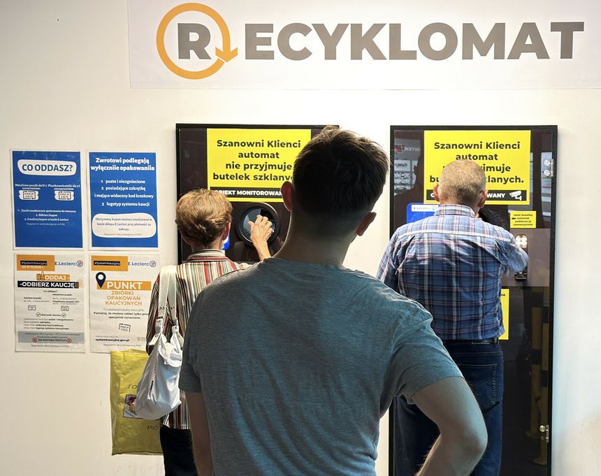 Trzy osoby korzystają z automatu do recyklingu, przystojąc przed wyświetlaczami informacyjnymi.