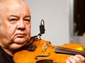 Mężczyzna z instrumentem smyczkowym, z mikrofonem, na tle szarej ściany.