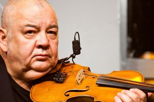 Mężczyzna z instrumentem smyczkowym, z mikrofonem, na tle szarej ściany.