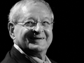Osoba w okularach, uśmiechająca się, ciemne tło, portret w odcieniach czerni i bieli.