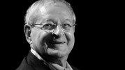Osoba w okularach, uśmiechająca się, ciemne tło, portret w odcieniach czerni i bieli.