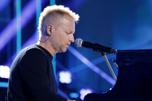 Mężczyzna przy pianinie, śpiewający do mikrofonu, w tle kolorowe oświetlenie sceny.