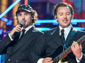 Na scenie dwóch mężczyzn w eleganckich garniturach, jeden śpiewa, drugi gra na gitarze.