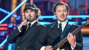Na scenie dwóch mężczyzn w eleganckich garniturach, jeden śpiewa, drugi gra na gitarze.