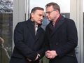 Dwaj mężczyźni w zimowych płaszczach, rozmawiający pod szklanymi drzwiami.