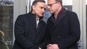 Dwaj mężczyźni w zimowych płaszczach, rozmawiający pod szklanymi drzwiami.