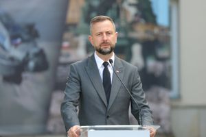 Na zdjęciu mężczyzna w garniturze przy mównicy, wyraża uwagę podczas przemówienia.