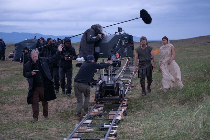 Na planie filmowym, ekipa techniczna ustawia kamerę na torach, w tle aktorzy w kostiumach.