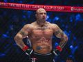 Piotr „Bestia” Piechowiak z tatuażami stoi w klatce MMA, ubrany w rękawice. Tło jasno oświetlone.