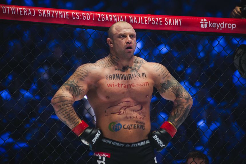 Piotr „Bestia” Piechowiak z tatuażami stoi w klatce MMA, ubrany w rękawice. Tło jasno oświetlone.