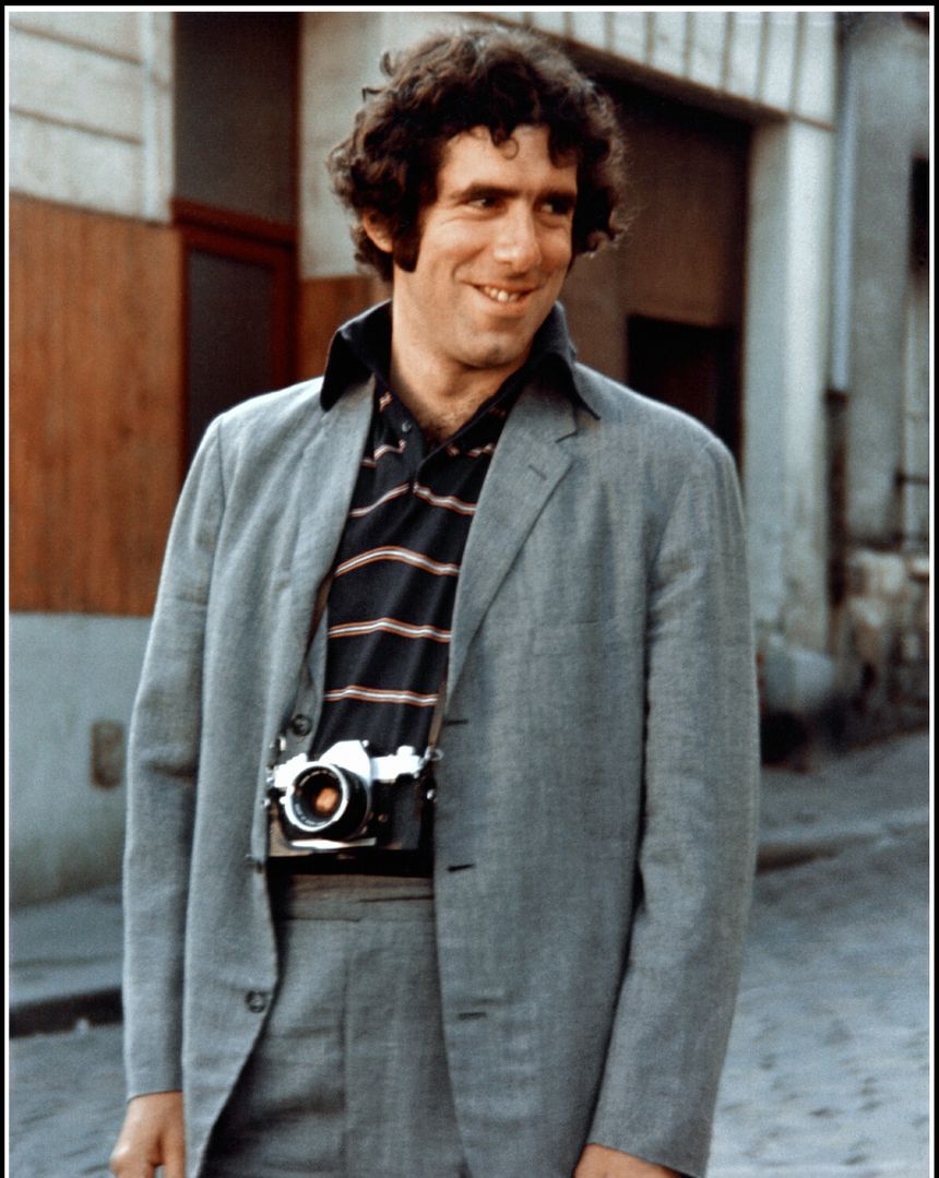 Mężczyzna w szarym garniturze z aparatem fotograficznym, uśmiechnięty na ulicy.