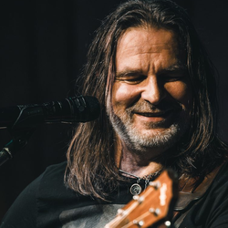Mężczyzna z długimi włosami gra na gitarze i śpiewa przy mikrofonie. Uśmiecha się.