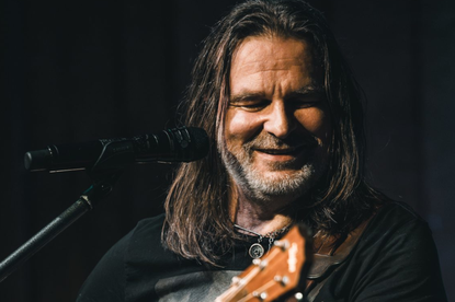 Mężczyzna z długimi włosami gra na gitarze i śpiewa przy mikrofonie. Uśmiecha się.