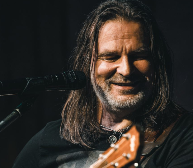 Mężczyzna z długimi włosami gra na gitarze i śpiewa przy mikrofonie. Uśmiecha się.