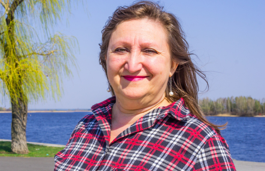 Kobieta w koszuli w kratę, uśmiechnięta, stoi nad brzegiem wody. W tle drzewa i błękitne niebo.