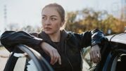 Kobieta w skórzanej kurtce opiera się o samochód, w tle widać świecące słońce i drzewa.