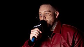 Michał Boxdel Baron w czerwonej kurtce trzyma mikrofon i mówi, stoi na ciemnym tle.