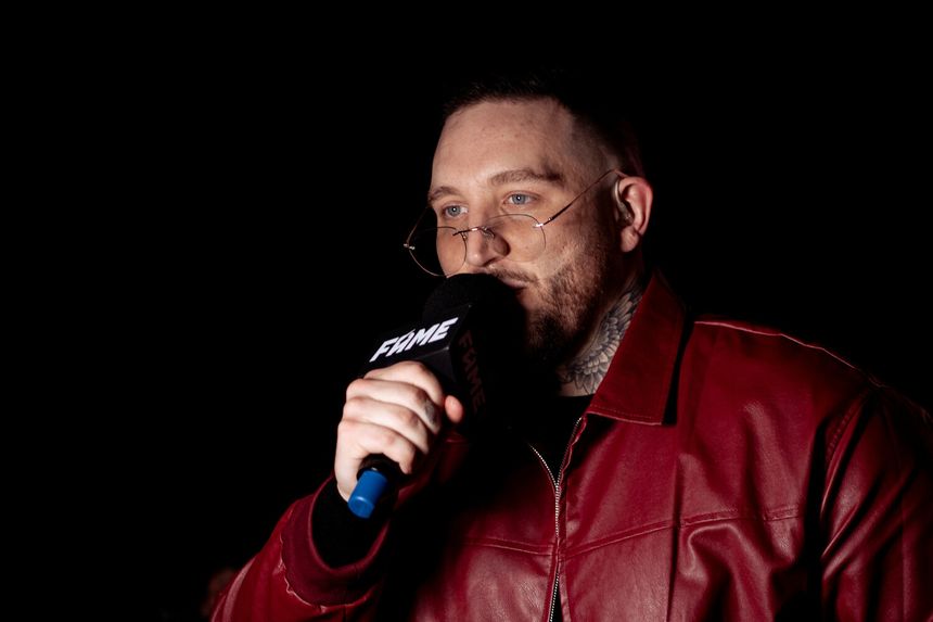 Michał Boxdel Baron w czerwonej kurtce trzyma mikrofon i mówi, stoi na ciemnym tle.