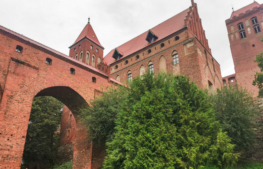 Zamek z czerwonej cegły otoczony zielenią, pod chmurami, z mostem w tle.