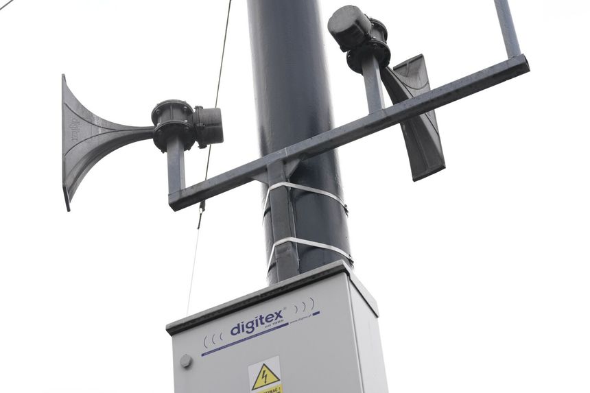 Na zdjęciu widać głośniki na metalowym słupie, obok obudowa elektryczna. Szary, pochmurny dzień.